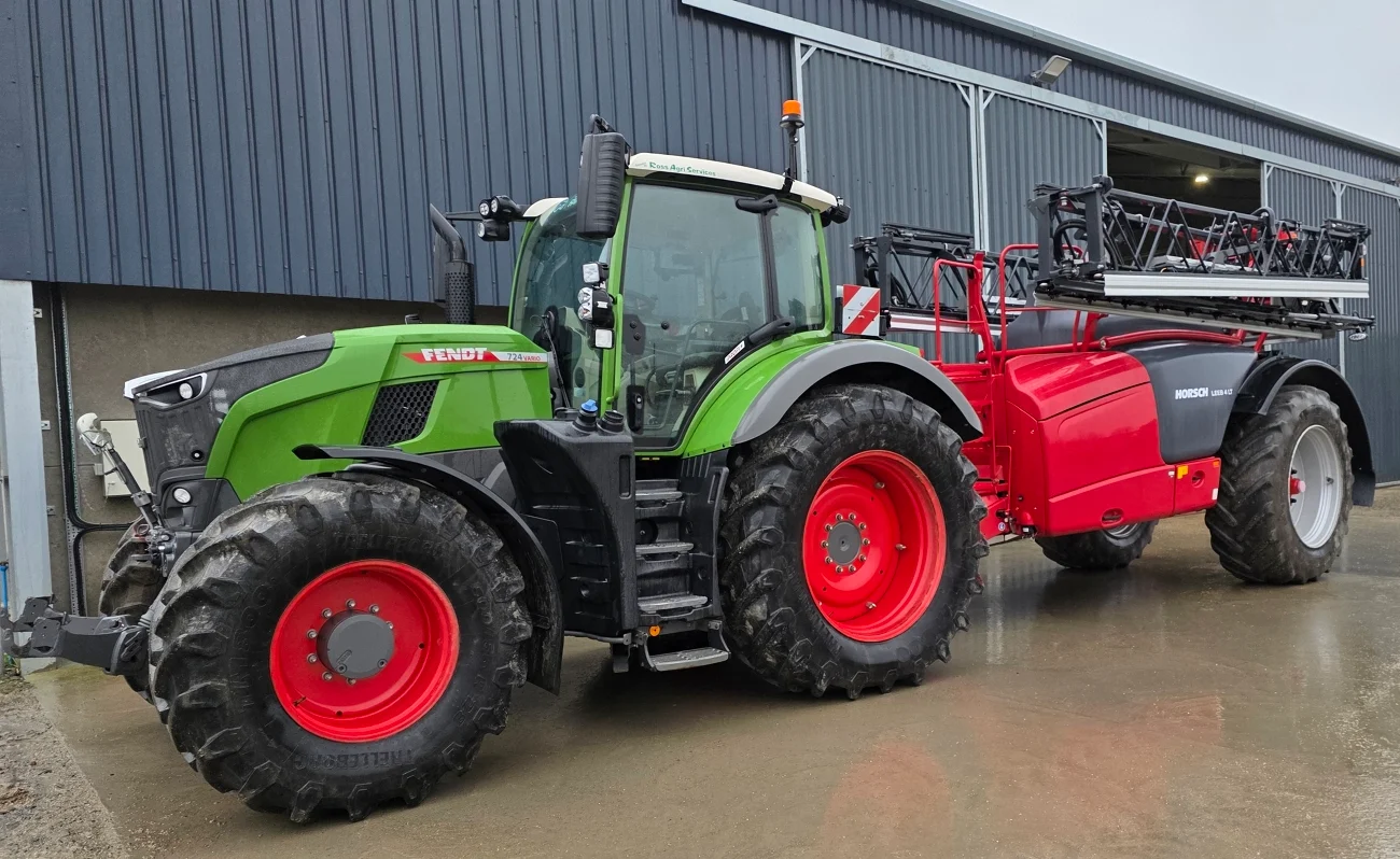 Fendt 724 Vario Gen7 mit roten Felgen und angebauter Horsch-Feldspritze vor einer Maschinenhalle